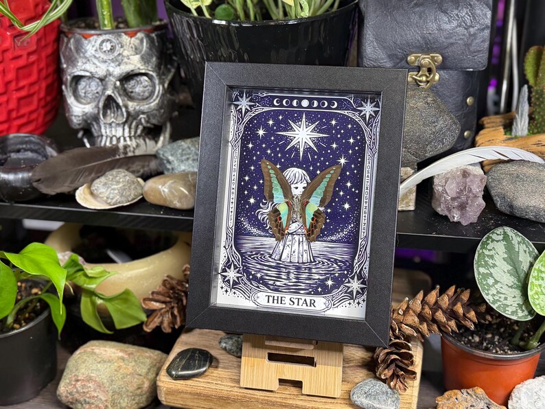 Op de afbeelding: Een ingelijste print van de tarotkaart "De Ster". Het kunstwerk toont een figuur met vlindervleugels die in het water staat onder een sterrenhemel. De zwarte lijst bevat de print, die op een houten standaard wordt getoond. Het thema is mystiek en hemels.