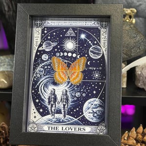May include: Framed artwork depicting a butterfly with orange and yellow wings, set against a cosmic background with planets, stars, and the text "THE LOVERS." The artwork is in a black frame.