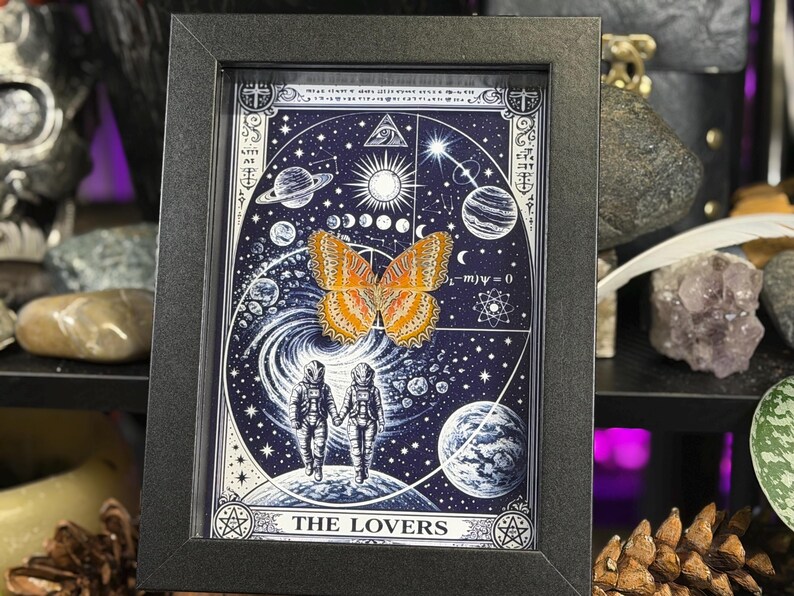 May include: Framed artwork depicting a butterfly with orange and yellow wings, set against a cosmic background with planets, stars, and the text "THE LOVERS." The artwork is in a black frame.