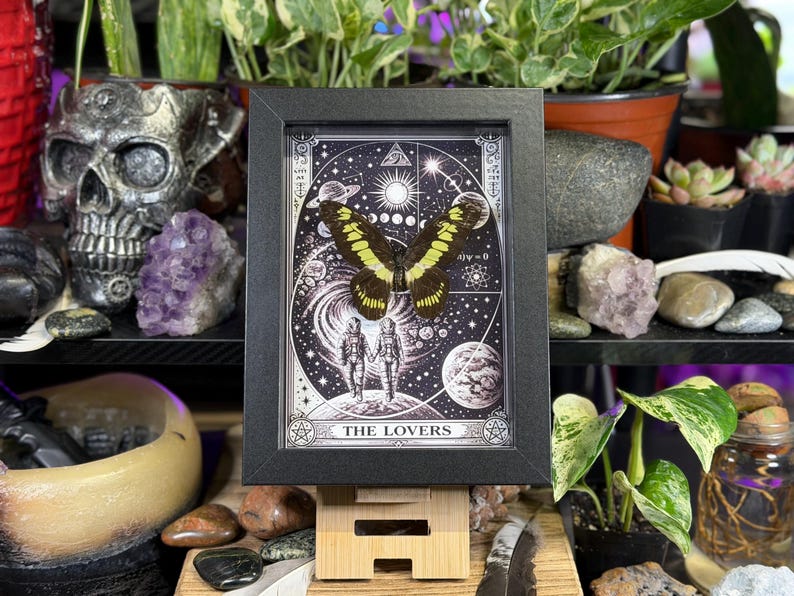 Puede incluir: Una obra de arte enmarcada que presenta una mariposa con alas amarillas y negras, sobre un fondo celestial con planetas, estrellas y el texto "THE LOVERS". La obra est&aacute; en un marco negro, exhibida sobre un soporte de madera, rodeada de plantas y objetos decorativos.