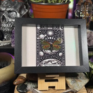 May include: A framed artwork depicting a butterfly with orange, green, and brown wings, set against a black and white celestial background. The artwork is labelled "The Universe" and is in a black frame.