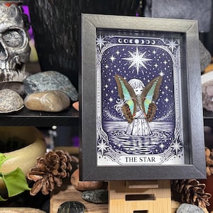 Op de afbeelding: Ingelijste kunst met een gedetailleerde illustratie van een figuur met vlindervleugels tegen een sterrenhemel. Het kunstwerk is voorzien van het opschrift "THE STAR" en is ingelijst in een zwarte lijst. De afbeelding heeft een mystiek en hemels thema.