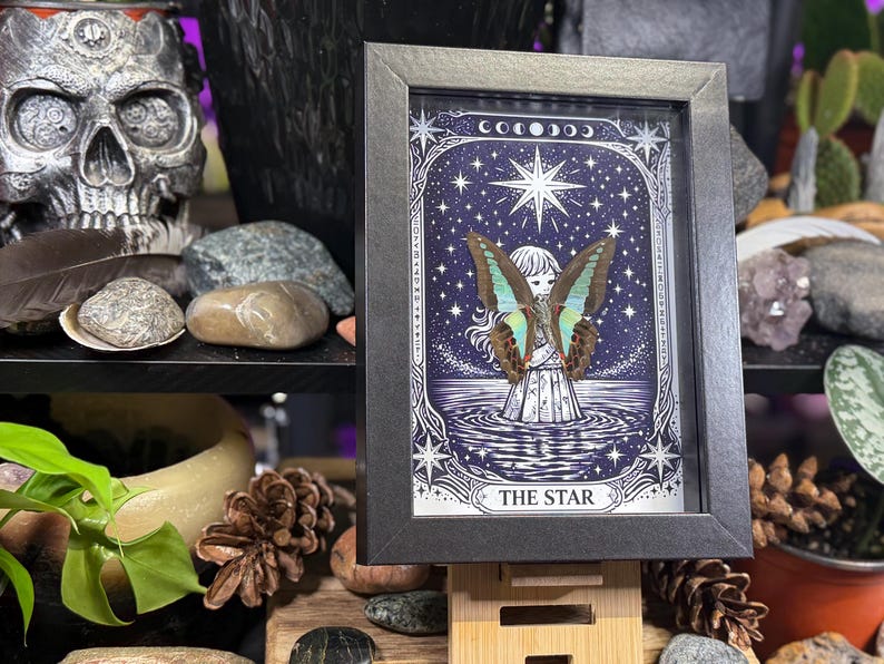 Op de afbeelding: Ingelijste kunst met een gedetailleerde illustratie van een figuur met vlindervleugels tegen een sterrenhemel. Het kunstwerk is voorzien van het opschrift "THE STAR" en is ingelijst in een zwarte lijst. De afbeelding heeft een mystiek en hemels thema.
