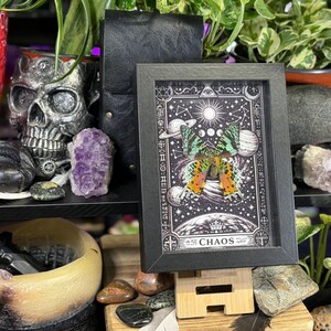 May include: A black framed print displaying a butterfly with orange, green, and black wings, set against a celestial background with planets, moons, and stars. The word "CHAOS" is at the bottom.