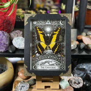 Peut inclure: Une &oelig;uvre encadr&eacute;e repr&eacute;sentant un papillon jaune et noir sur un fond c&eacute;leste. Le cadre noir a des coins dor&eacute;s. L'&oelig;uvre est intitul&eacute;e "THE UNIVERSE" en bas.