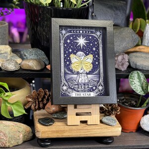 May include: A framed tarot card print of "The Star" on a wooden easel. The artwork features a figure with butterfly wings, set against a starry night sky. The frame is black, and the print has a blue and white colour scheme.