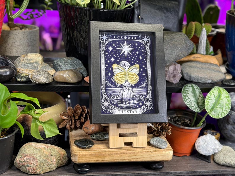 May include: A framed tarot card print of "The Star" on a wooden easel. The artwork features a figure with butterfly wings, set against a starry night sky. The frame is black, and the print has a blue and white colour scheme.