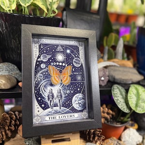 May include: Black framed artwork with a celestial design, planets, stars, and two figures holding hands. An orange butterfly is centered on the image. The words "THE LOVERS" are at the bottom. The artwork is displayed on a wooden surface.