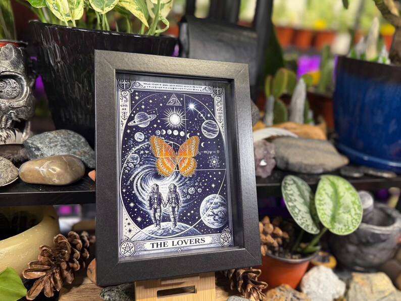 May include: Black framed artwork with a celestial design, planets, stars, and two figures holding hands. An orange butterfly is centered on the image. The words "THE LOVERS" are at the bottom. The artwork is displayed on a wooden surface.