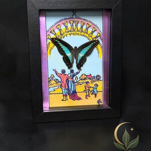Puede incluir: Un marco negro tipo caja que contiene una ilustraci&oacute;n de la carta del tarot Diez de Copas con una mariposa central de alas negras y turquesas. La obra muestra a dos figuras con los brazos levantados hacia un arco&iacute;ris de diez copas doradas.