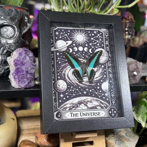 Op de afbeelding: Zwartgelijste kunstdruk met een vlinder met turquoise en zwarte vleugels, tegen een zwart-witte hemelachtergrond. De print bevat planeten, sterren en de tekst "The Universe".