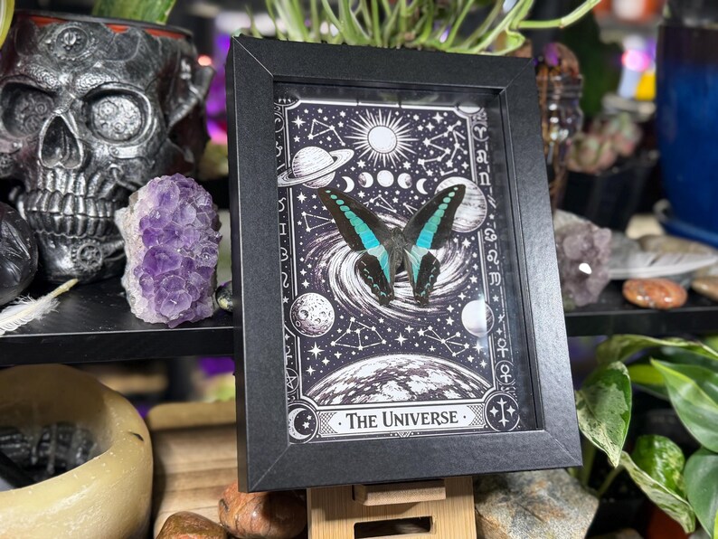 Op de afbeelding: Zwartgelijste kunstdruk met een vlinder met turquoise en zwarte vleugels, tegen een zwart-witte hemelachtergrond. De print bevat planeten, sterren en de tekst "The Universe".
