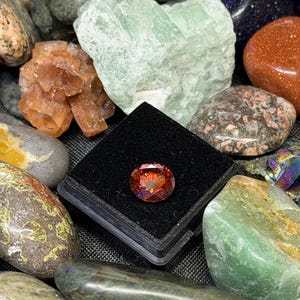 Puede incluir: Una colecci&oacute;n de varias piedras preciosas y cristales. Una gema facetada, redonda, de color naranja rojizo se muestra en una caja negra. Alrededor de la gema hay otras piedras en tonos verdes, marrones, naranjas y grises.