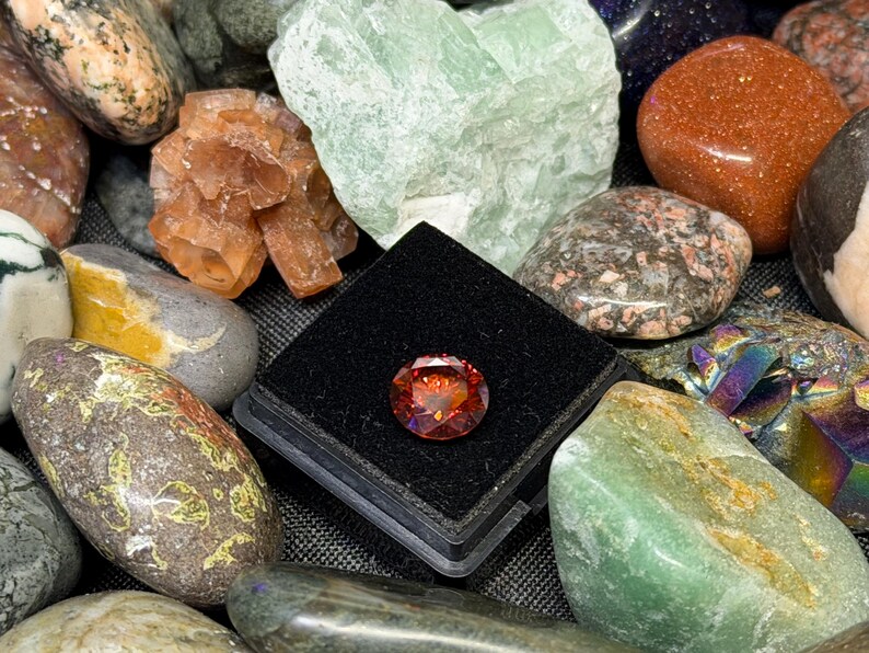Puede incluir: Una colecci&oacute;n de varias piedras preciosas y cristales. Una gema facetada, redonda, de color naranja rojizo se muestra en una caja negra. Alrededor de la gema hay otras piedras en tonos verdes, marrones, naranjas y grises.