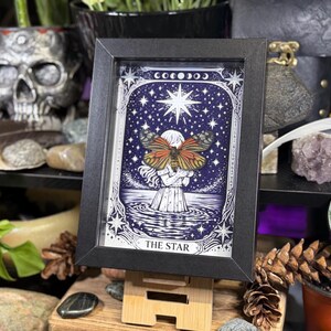 May include: Framed art print of "The Star" tarot card. The image features a woman holding a butterfly with a starry night sky background. The print is in a black frame and displayed on a wooden easel. The overall color scheme is blue, white, and orange.