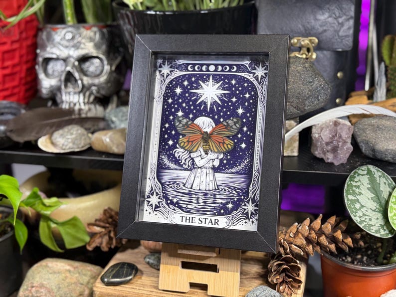 May include: Framed art print of "The Star" tarot card. The image features a woman holding a butterfly with a starry night sky background. The print is in a black frame and displayed on a wooden easel. The overall color scheme is blue, white, and orange.