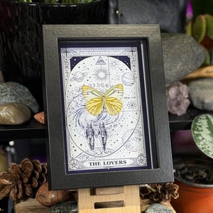 K&ouml;nnte beinhalten: Gerahmtes Kunstwerk mit einem gelben Schmetterling &uuml;ber einem Himmelsdesign mit Planeten, Sternen und dem Schriftzug "THE LOVERS". Der schwarze Rahmen h&auml;lt das Kunstwerk, das auf einer Holzstaffelei steht. Das Kunstwerk ist von Steinen und Pflanzen umgeben.