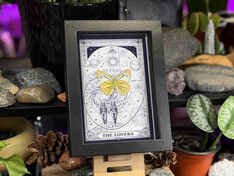 K&ouml;nnte beinhalten: Gerahmtes Kunstwerk mit einem gelben Schmetterling &uuml;ber einem Himmelsdesign mit Planeten, Sternen und dem Schriftzug "THE LOVERS". Der schwarze Rahmen h&auml;lt das Kunstwerk, das auf einer Holzstaffelei steht. Das Kunstwerk ist von Steinen und Pflanzen umgeben.