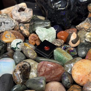 Puede incluir: Una colecci&oacute;n de diversas piedras preciosas pulidas y rocas en bruto en una variedad de colores, incluyendo verde, naranja, marr&oacute;n y gris. Una peque&ntilde;a caja negra cuadrada contiene una sola piedra preciosa roja. Las piedras var&iacute;an en tama&ntilde;o y forma.