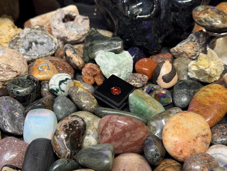 Puede incluir: Una colecci&oacute;n de diversas piedras preciosas pulidas y rocas en bruto en una variedad de colores, incluyendo verde, naranja, marr&oacute;n y gris. Una peque&ntilde;a caja negra cuadrada contiene una sola piedra preciosa roja. Las piedras var&iacute;an en tama&ntilde;o y forma.