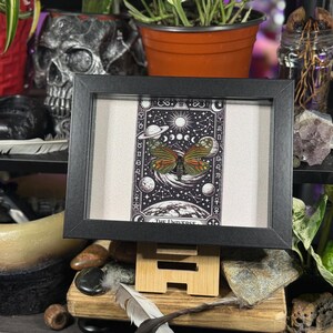 May include: A framed print with a butterfly design, showcasing orange and green wings against a backdrop of planets, moons, and stars. The black framed print is displayed on a wooden stand.