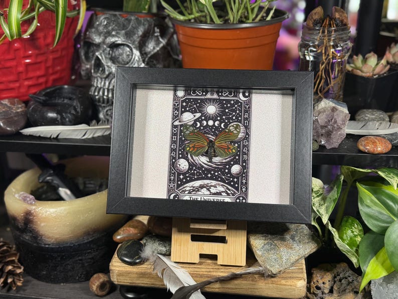 May include: A framed print with a butterfly design, showcasing orange and green wings against a backdrop of planets, moons, and stars. The black framed print is displayed on a wooden stand.