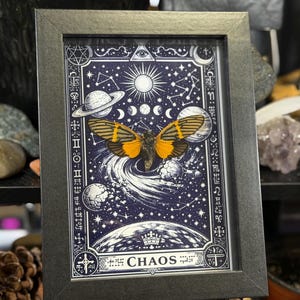May include: Framed artwork featuring a celestial design with a central image of a butterfly with orange and black wings. The artwork is set against a dark blue background with planets, stars, and the word "CHAOS" at the bottom.