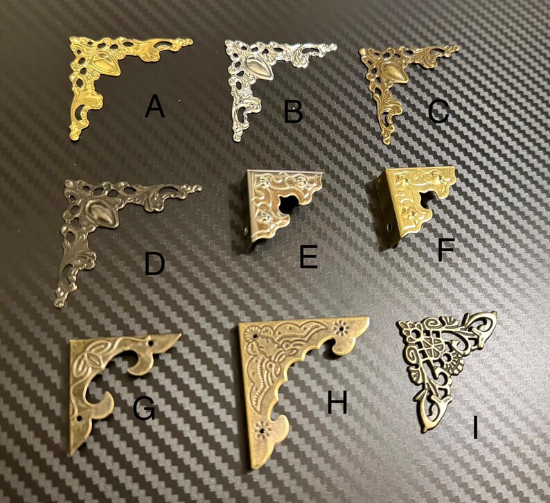 Puede incluir: Nueve escuadras de esquina decorativas de metal en varios acabados, incluyendo oro, plata y bronce. Cada escuadra presenta un dise&ntilde;o ornamentado de inspiraci&oacute;n vintage con detalles intrincados. Las escuadras est&aacute;n dispuestas sobre una superficie oscura y texturizada.