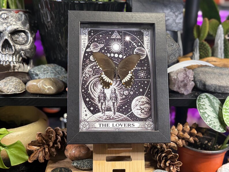 Puede incluir: Una obra de arte enmarcada con una mariposa de alas marrones oscuras y marcas blancas. La obra tiene un marco negro y representa una escena celestial con planetas, estrellas y las palabras "THE LOVERS". El tema general es m&iacute;stico y esot&eacute;rico.