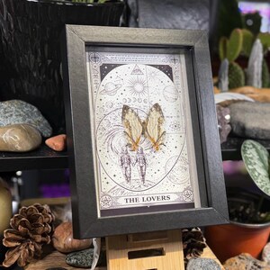 May include: A framed print featuring a tarot card design. The artwork depicts two figures with butterfly wings, set against a celestial background with planets and stars. The text "THE LOVERS" is at the bottom, framed in a black wooden frame.