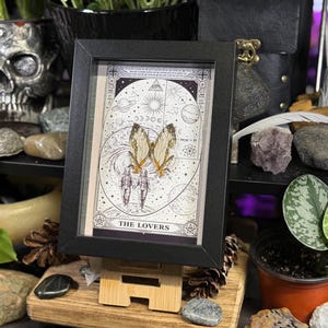 May include: A framed artwork featuring a tarot card design with the text "THE LOVERS". The artwork includes illustrations of butterflies, celestial bodies, and figures. The frame is black, and the artwork is displayed on a wooden stand.