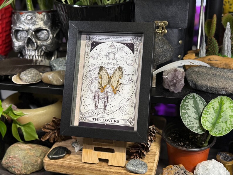 May include: A framed artwork featuring a tarot card design with the text "THE LOVERS". The artwork includes illustrations of butterflies, celestial bodies, and figures. The frame is black, and the artwork is displayed on a wooden stand.