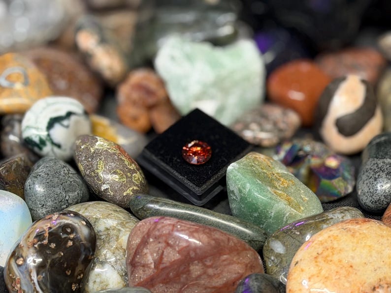 Puede incluir: Una colecci&oacute;n de piedras preciosas y rocas pulidas de varios colores, incluyendo verde, marr&oacute;n y naranja. Una peque&ntilde;a gema naranja facetada se exhibe sobre una superficie de terciopelo negro, rodeada de otras piedras. Primer plano.