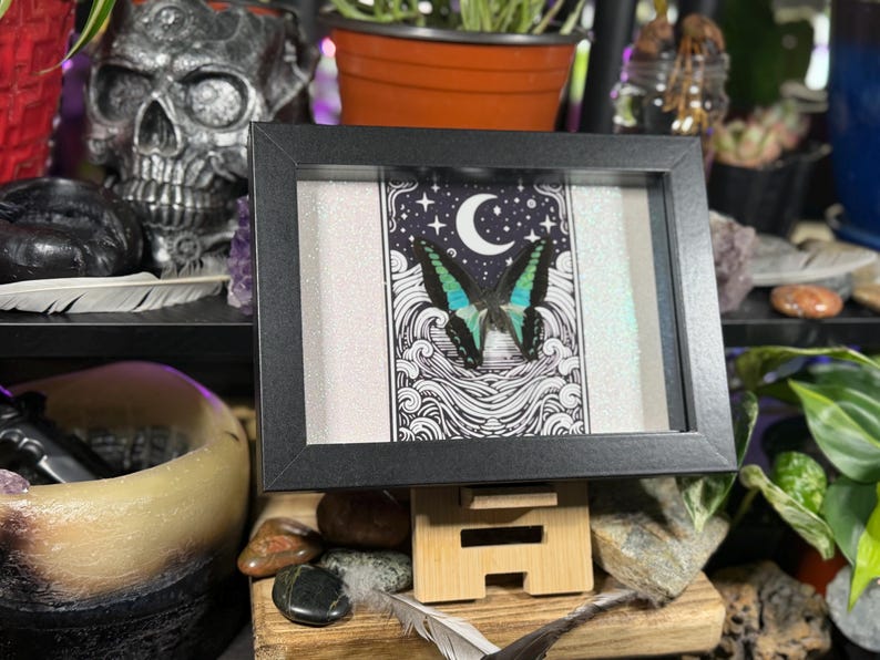 May include: A framed artwork featuring a butterfly with blue and green wings, set against a black and white celestial background with a crescent moon and stars. The artwork is in a black frame and displayed on a wooden stand.
