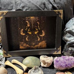 May include: A black framed art piece with gold corner accents, featuring a celestial design with moons, stars, and a sun. The artwork is surrounded by crystals, bones, and a skull, creating a mystical aesthetic.