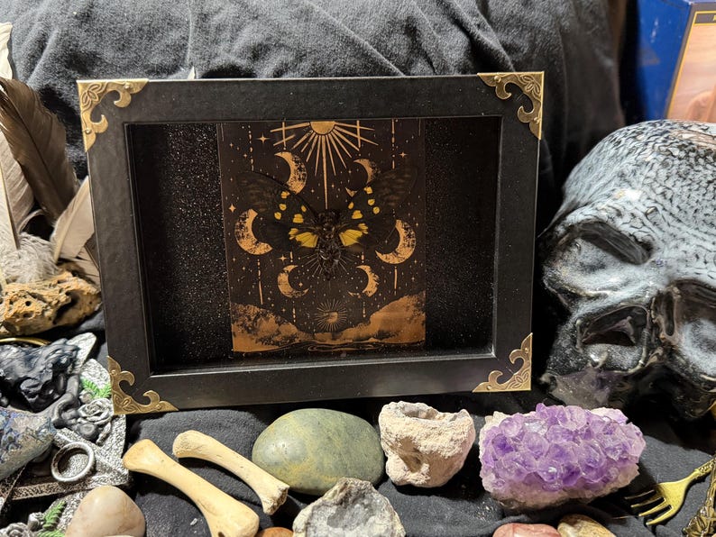 May include: A black framed art piece with gold corner accents, featuring a celestial design with moons, stars, and a sun. The artwork is surrounded by crystals, bones, and a skull, creating a mystical aesthetic.