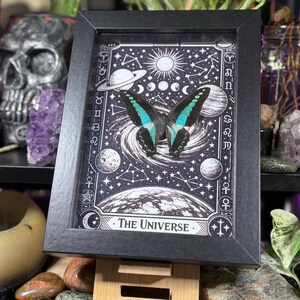 Op de afbeelding: Een ingelijst kunstwerk met een vlinder met zwarte en turquoise vleugels, tegen een zwart-witte hemelse achtergrond. Het kunstwerk bevat planeten, sterren en de tekst "The Universe". De lijst is zwart.