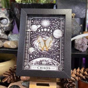 May include: A framed artwork with a black and white celestial design, including planets, stars, and the word "CHAOS". The artwork features a butterfly with delicate wings. The frame is black and the artwork is displayed on a wooden easel.