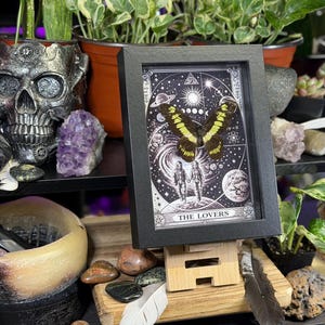 Puede incluir: Una impresi&oacute;n enmarcada en negro de la carta del tarot "Los Amantes". La obra de arte presenta una mariposa con alas amarillas y negras sobre un fondo estrellado con elementos celestiales.