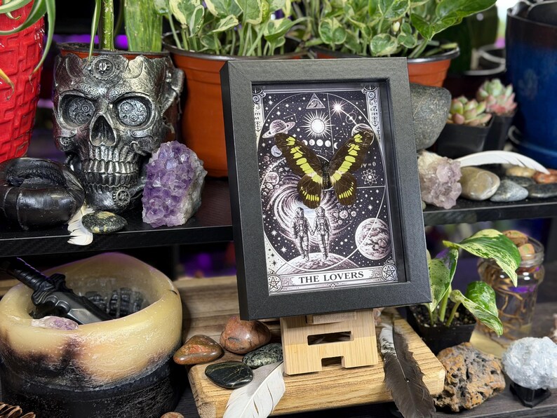 Puede incluir: Una impresi&oacute;n enmarcada en negro de la carta del tarot "Los Amantes". La obra de arte presenta una mariposa con alas amarillas y negras sobre un fondo estrellado con elementos celestiales.