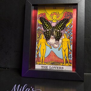 May include: A framed artwork featuring The Lovers tarot card with a large, dark butterfly with green spots mounted over the central figure. The card displays two yellow figures, a sun, and a mountain landscape. The frame is black with red accents.