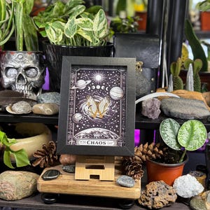May include: A framed art print with a celestial design, featuring planets, moons, and stars, with the word "CHAOS" at the bottom. The print is displayed on a wooden easel, surrounded by plants, rocks, and decorative items.