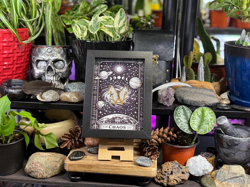 May include: A framed art print with a celestial design, featuring planets, moons, and stars, with the word "CHAOS" at the bottom. The print is displayed on a wooden easel, surrounded by plants, rocks, and decorative items.