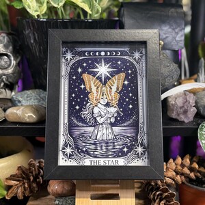 May include: Black framed print of "The Star" tarot card. The artwork depicts a woman with butterfly wings, standing in water under a starry sky. The print has a black frame and is displayed on a wooden stand.