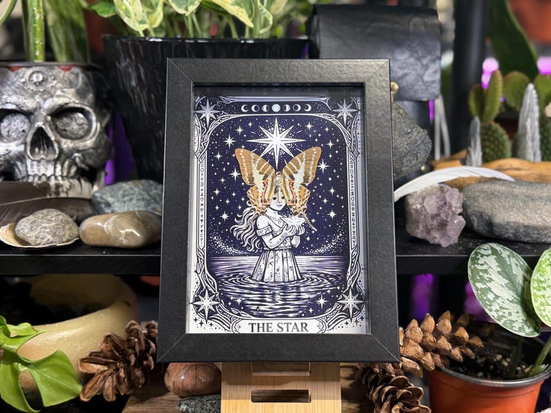 May include: Black framed print of "The Star" tarot card. The artwork depicts a woman with butterfly wings, standing in water under a starry sky. The print has a black frame and is displayed on a wooden stand.