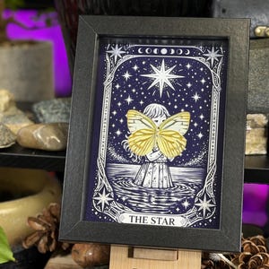 May include: Framed art print depicting a celestial scene with a figure holding a yellow butterfly. The artwork is set against a dark blue starry background, with the words "THE STAR" at the bottom. The frame is black.