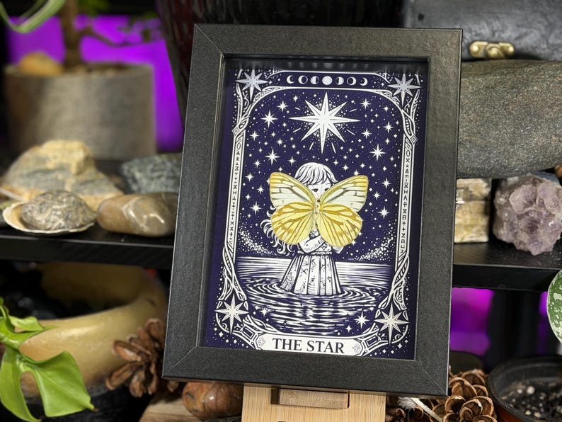 May include: Framed art print depicting a celestial scene with a figure holding a yellow butterfly. The artwork is set against a dark blue starry background, with the words "THE STAR" at the bottom. The frame is black.