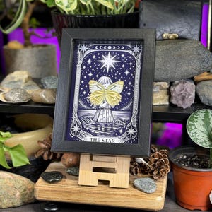 May include: A framed print of "The Star" tarot card. The artwork depicts a figure with butterfly wings against a starry night sky. The print is in a black frame and displayed on a wooden easel, surrounded by plants and decorative stones.