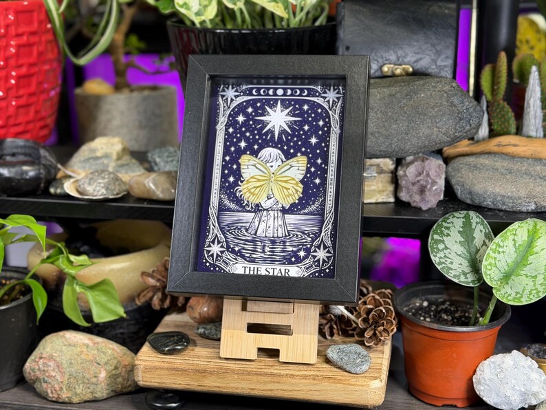 May include: A framed print of "The Star" tarot card. The artwork depicts a figure with butterfly wings against a starry night sky. The print is in a black frame and displayed on a wooden easel, surrounded by plants and decorative stones.