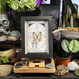 May include: A framed tarot card print of "The Lovers" with a black frame, displayed on a wooden easel. The card features a detailed illustration of two figures with butterfly wings, set against a celestial background. The image is surrounded by various decorative items.